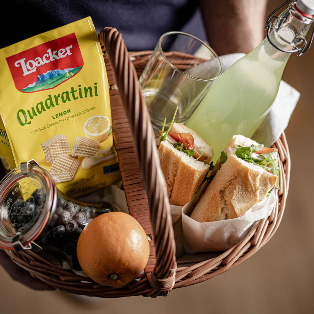 Quadratini Lemon, Crème-Filled Wafer Cookies, 8.82 oz. Bag in a basket with sandwiches, orange, blueberries in a jar, soda and glass.